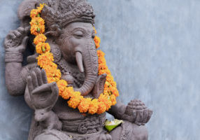Ganesha sitting in meditating yoga pose in front of hindu temple. Decorated for religious festival by orange flowers garland, ceremonial offering. Balinese travel background. Bali island art, culture.